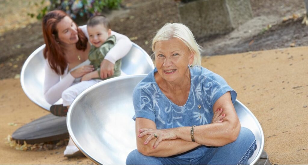 Eine Familiengesundheitspatin sitzt auf einem Spielgerät auf einem Spieltplatz. Im Hintergrund sieht man eine Mutter mit ihrem Kind.